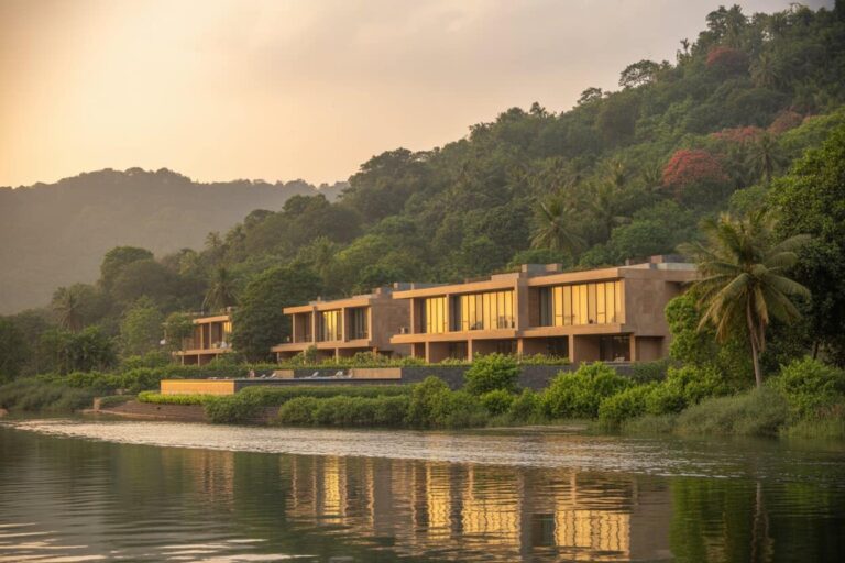 Row of contemporary beige villas along a calm river at sunset, surrounded by lush green hills and palm trees.
