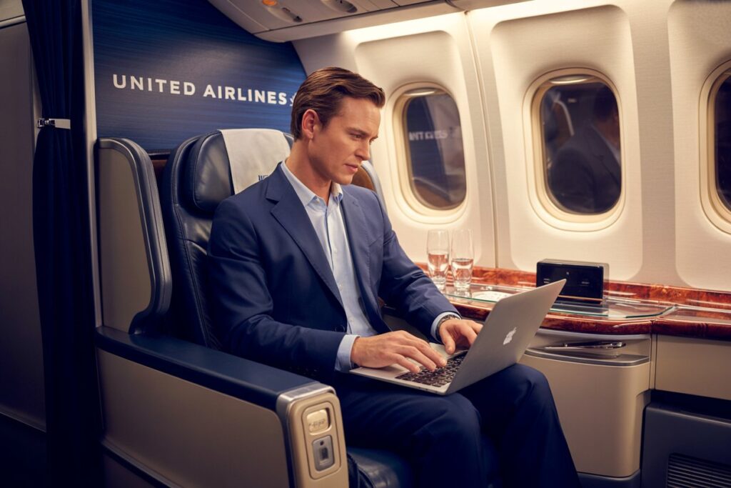 Business traveler in a blue suit typing on a laptop in a United Airlines first-class cabin, with window seats nearby.