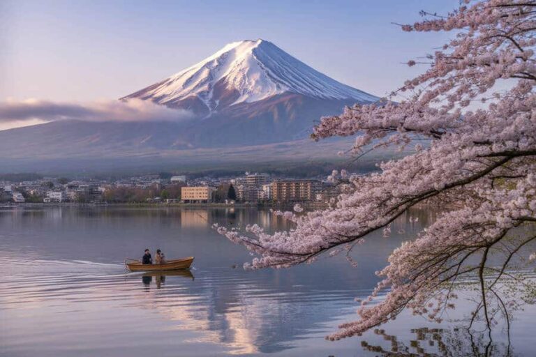 Two people row a small boat on a calm lake with Mount Fuji in the background and pink cherry blossoms along the shore on the right side.