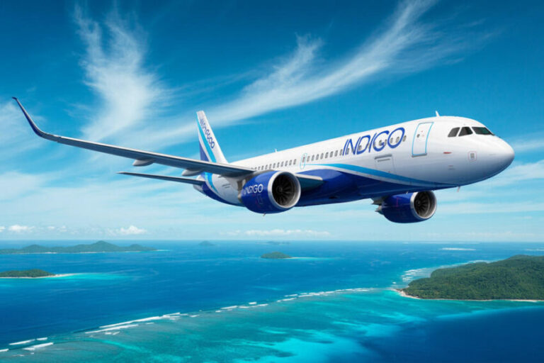 Indigo passenger plane in white and blue livery flying over a turquoise tropical ocean with small green islands below.