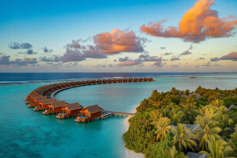 Aerial view of a curved row of overwater bungalows extending from a tropical island into turquoise lagoon under a sunset sky, with palm trees on the shore.