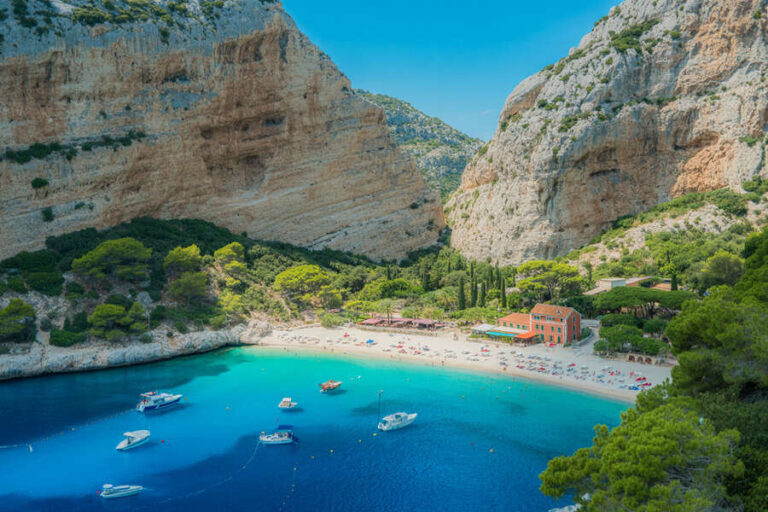 Aerial view of a turquoise cove with a sandy beach, moored boats, and a red building among green trees at the foot of tall cliffs.