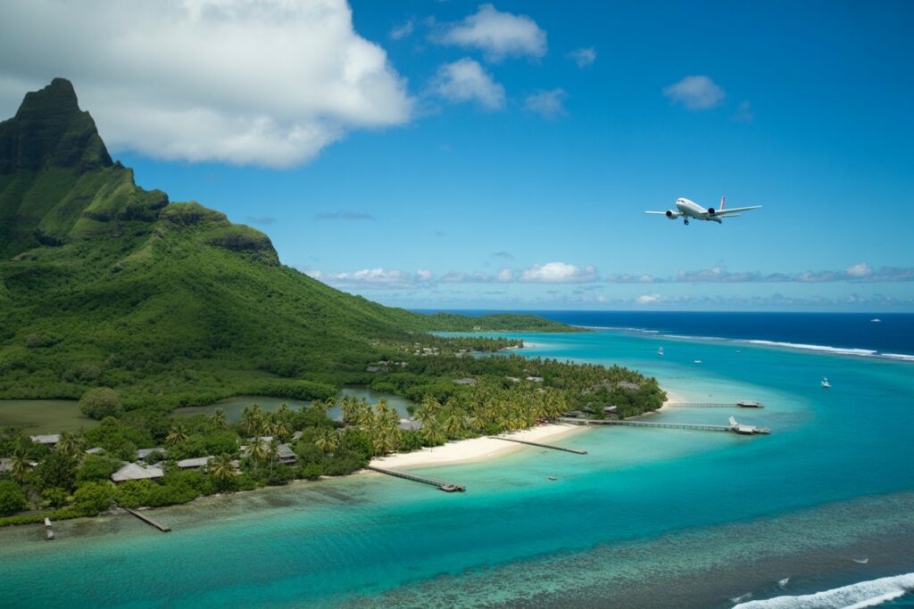 Tropical island with a green mountain, white-sand beaches, and turquoise water; an airplane in the blue sky.