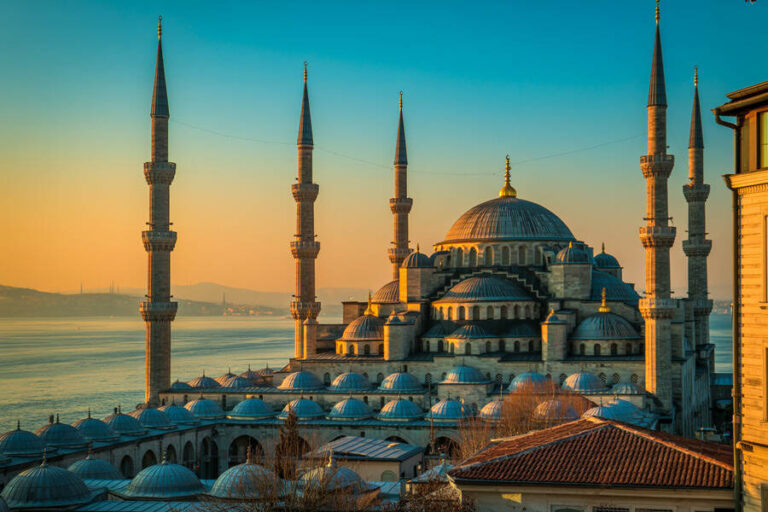 Sunset casts warm light on a grand Ottoman mosque with multiple minarets and domed roofs by the water.