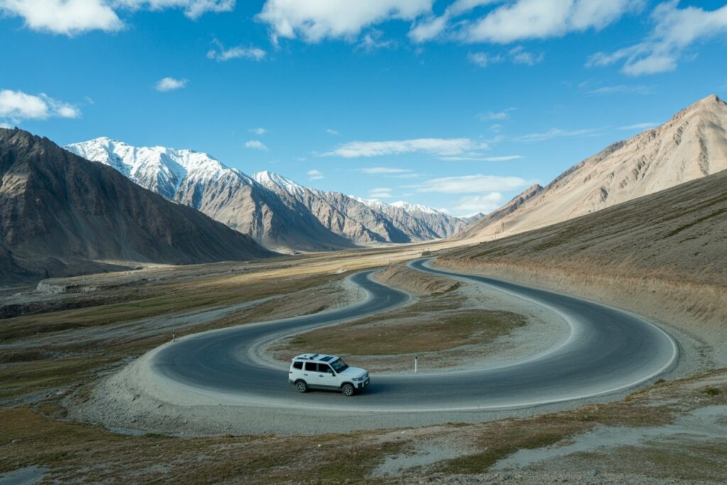 White SUV navigates a sharp hairpin turn on a winding mountain road under a blue sky with snow-capped peaks in the background.