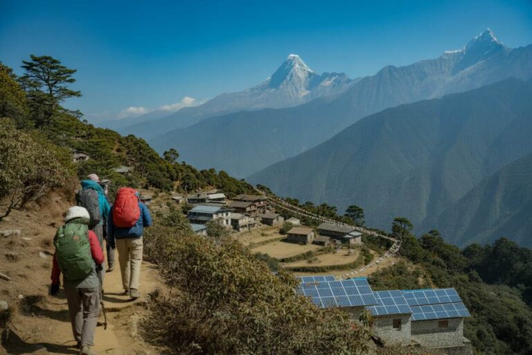 Hikers with large backpacks walk a dirt mountain path along a hillside village, with solar panels on a building and snow-capped peaks in the distance.