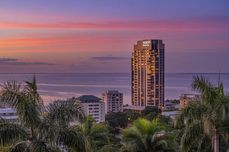 Sunset over a coastal city with a tall VOCO hotel tower reflecting golden light by the sea.