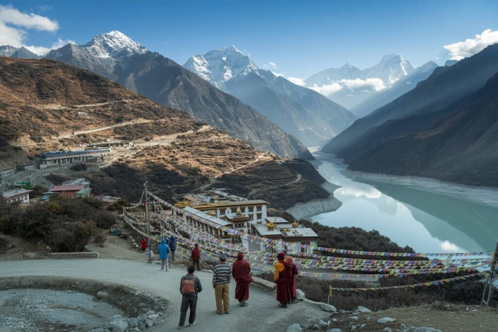 Terraced hillsides, prayer flags, and a winding river in a Himalayan valley with hikers on a dirt path and a small village nearby.