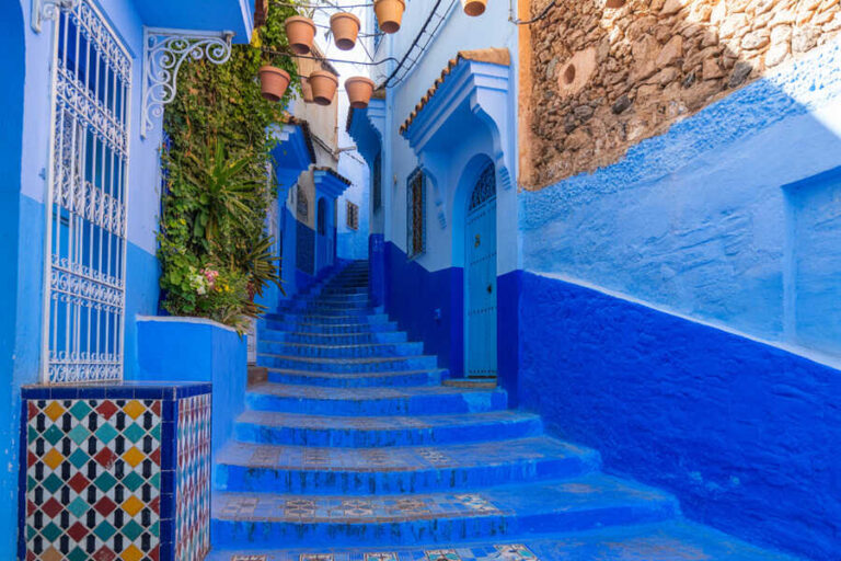 Blue-painted narrow alley with a stairway, potted plants, and decorative tiles at the left in a Moroccan town.