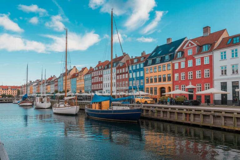 Colorful row of townhouses along a canal with moored sailboats and a wooden pier under a bright blue sky.