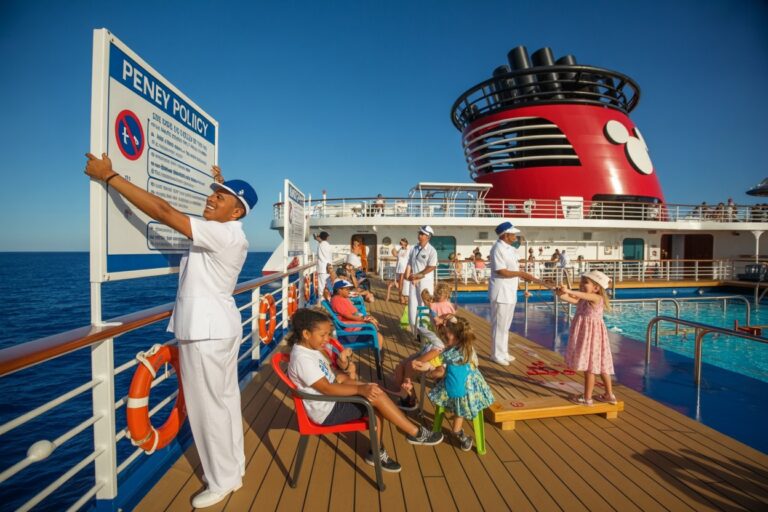 Crew members in white uniforms supervise passengers and children on a sunny cruise ship deck with a large poster and a red funnel in the background.