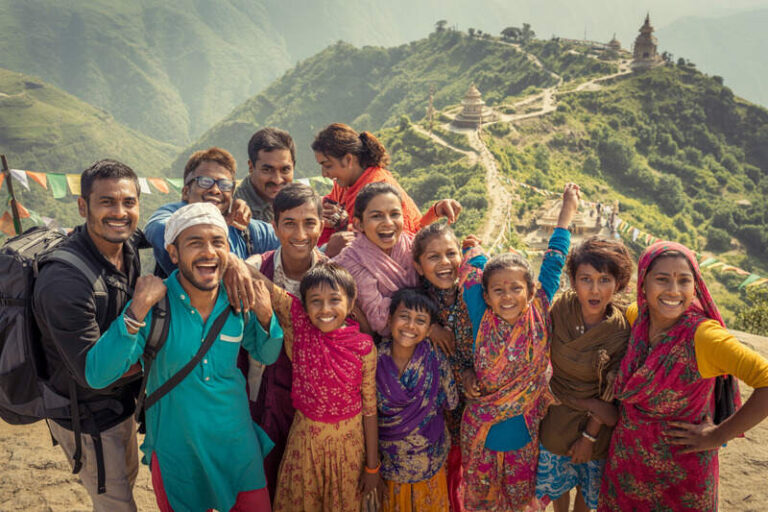 Smiling group of trekkers posing for a photo on a hillside trail in Nepal, with prayer flags and distant pagoda structures in the mountains behind them.