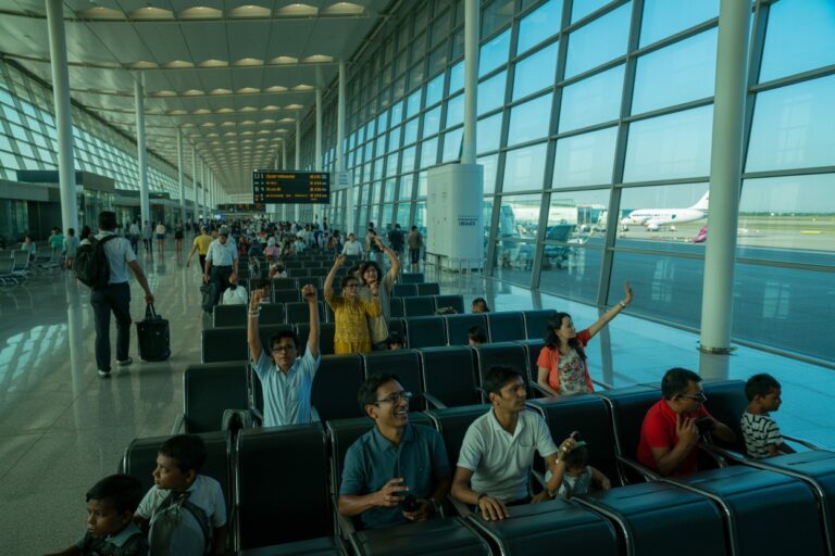 Passengers wait in an airport departure hall with large glass walls; some raise their hands as others sit and chat, planes outside visible through the windows.