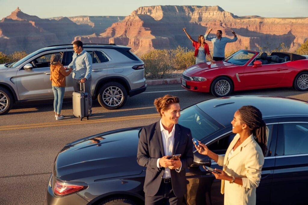 Group of four adults at a scenic canyon overlook, with parked cars and a red convertible; two are chatting near a black car in the foreground.