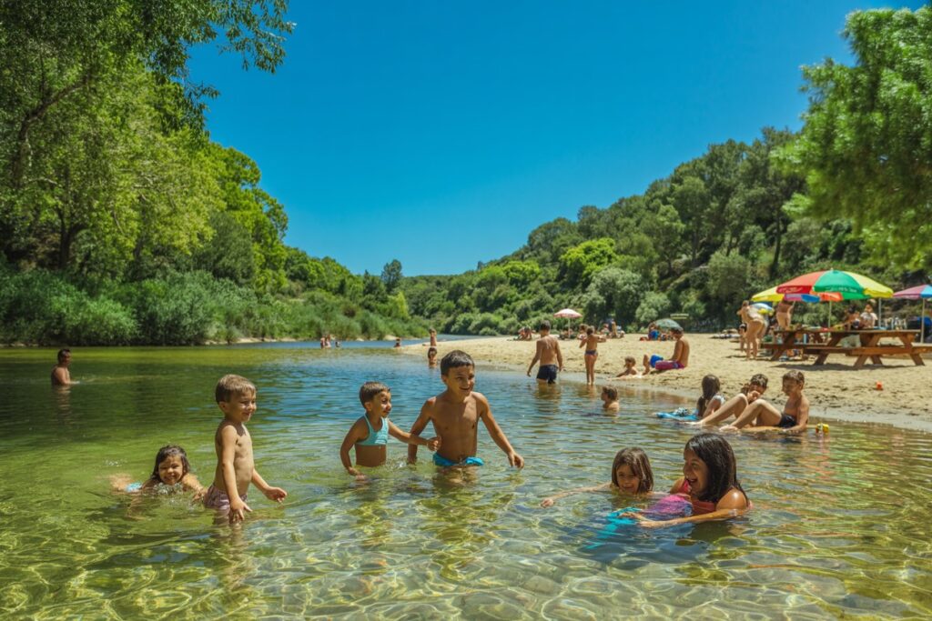 Kids wading and playing in a shallow, clear river at a sunny beach with a forest backdrop and people on the sandy shore.