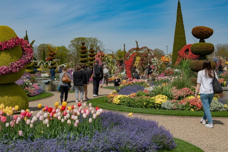 Visitors walk along a gravel path through a formal garden with colorful flower beds and sculpted topiary on a sunny day.