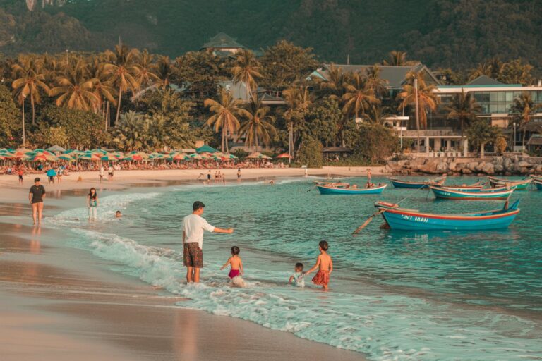 Families play in shallow surf at a sunny tropical beach with colorful umbrellas and palm trees in the background, boats anchored nearby.