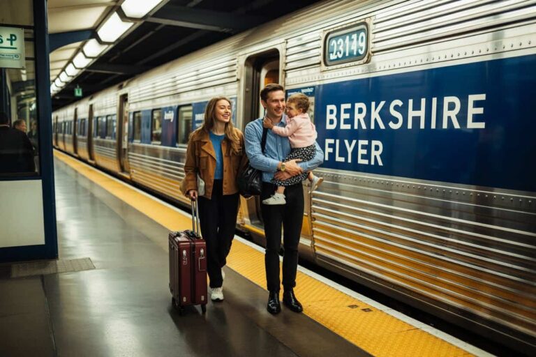 Family at a train platform beside a Berkshire Flyer train; father holds a child while mother stands with a rolling suitcase, smiling in conversation.