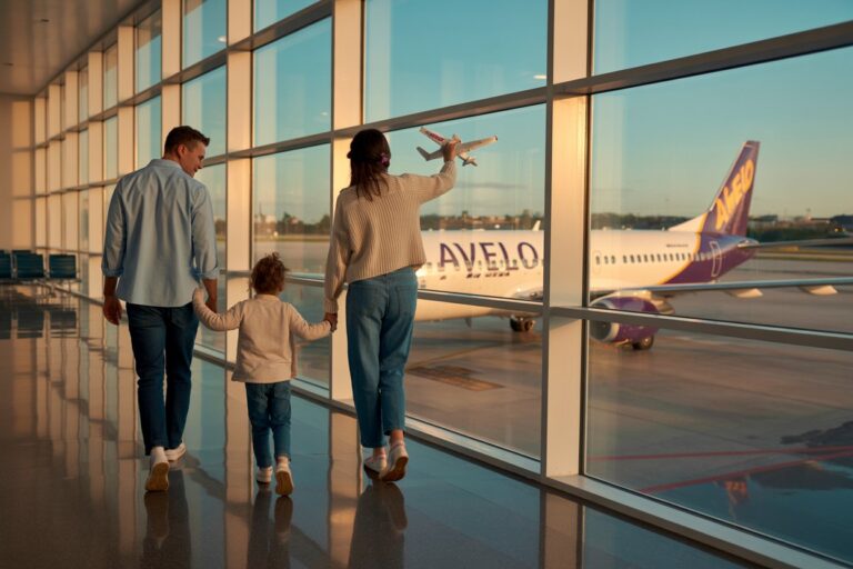 A family of three walks through a sunny airport terminal, holding hands and watching a white Avelo airplane outside the glass wall; the mother waves a toy plane as they pass by.