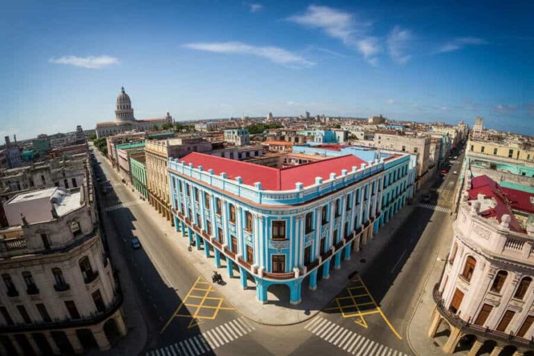 Aerial view of a bright blue historic building at a street intersection in Havana, with the Capitol dome visible in the distance on a clear day