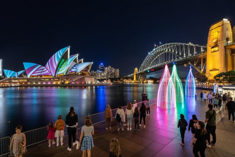 Night view of Sydney Harbour: illuminated Opera House with colorful sails, the Harbour Bridge, and a lit waterfront promenade with people and neon light installations along the water.