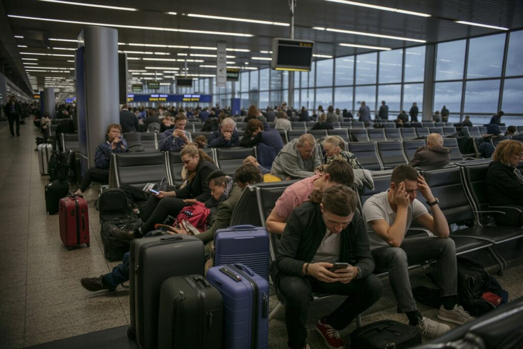 Airport waiting area with passengers seated among rolling suitcases and bags; some are using their phones.