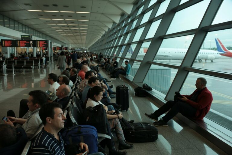 Busy airport terminal with rows of seated passengers and luggage, looking toward large angled windows with planes on the tarmac outside