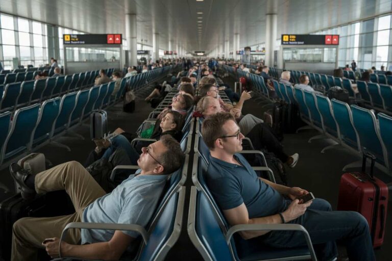 Crowded airport gate lounge with travelers seated in blue chairs and rolling suitcases nearby.