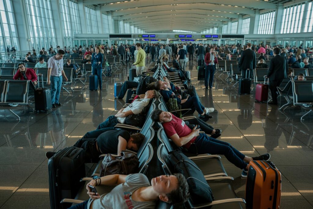 Passengers resting in a long row of seats at a busy airport terminal, with suitcases nearby.