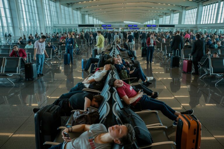 Passengers resting in a long row of seats at a busy airport terminal, with suitcases nearby.