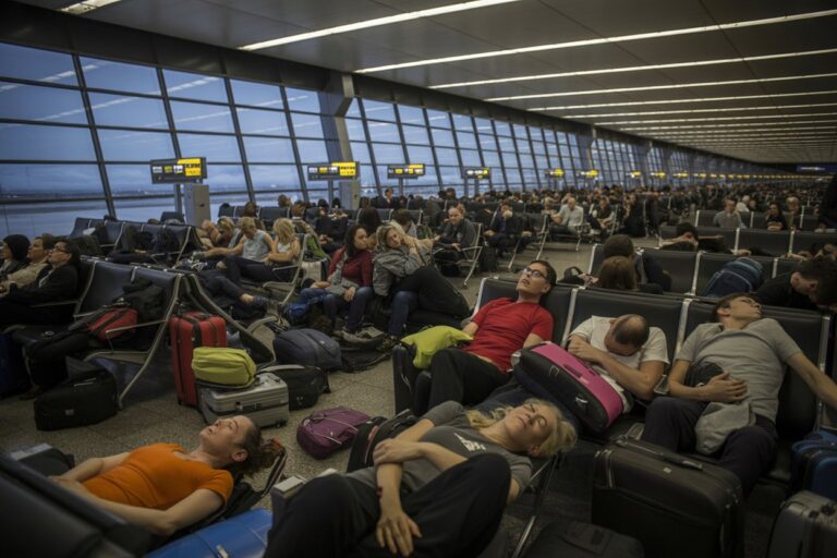 Busy airport terminal with many travelers sitting and reclining, luggage at their feet.