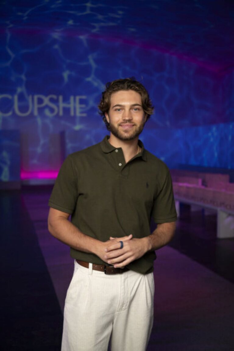 Man standing in a dim studio against a blue patterned background, wearing an olive polo shirt and white pants, hands clasped.