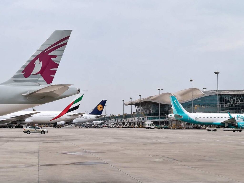 Several commercial airplanes parked at gates on a large airport tarmac, with a curved-roof terminal in the background and a turquoise aircraft on the right.