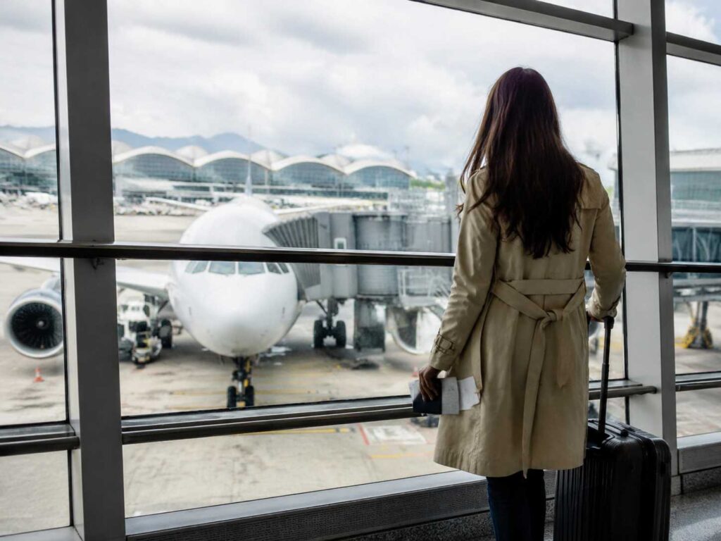 Back view of a woman in a beige trench coat standing with a rolling suitcase, looking out at a parked airplane through airport gates, holding boarding documents in one hand.