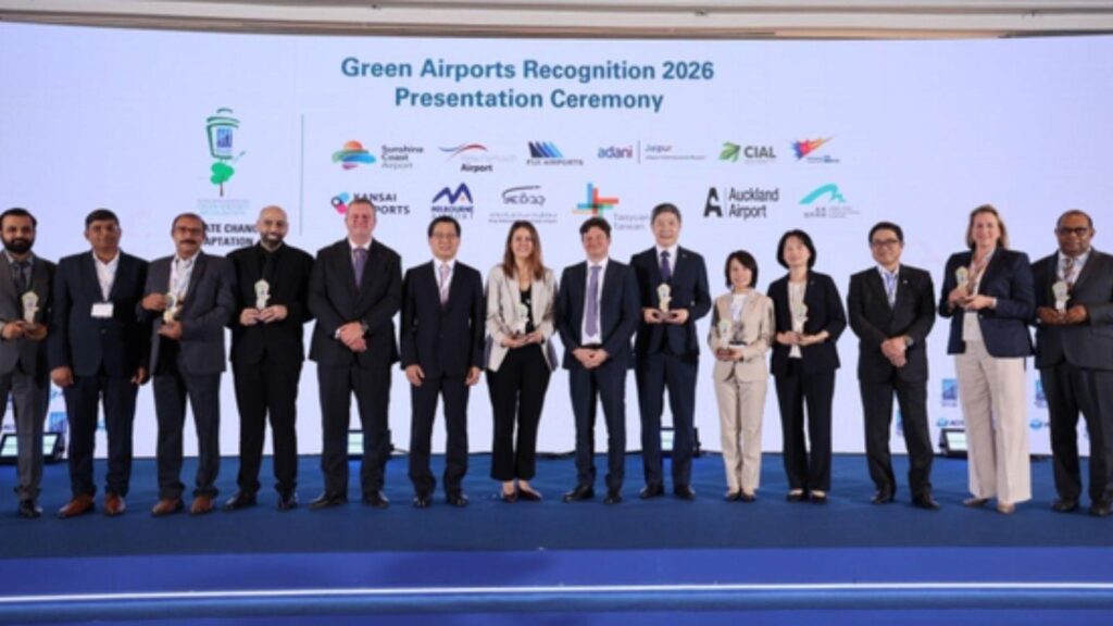 Group of award recipients in suits on a stage, each holding a trophy, at the Green Airports Recognition 2026 ceremony with a logos backdrop behind them.