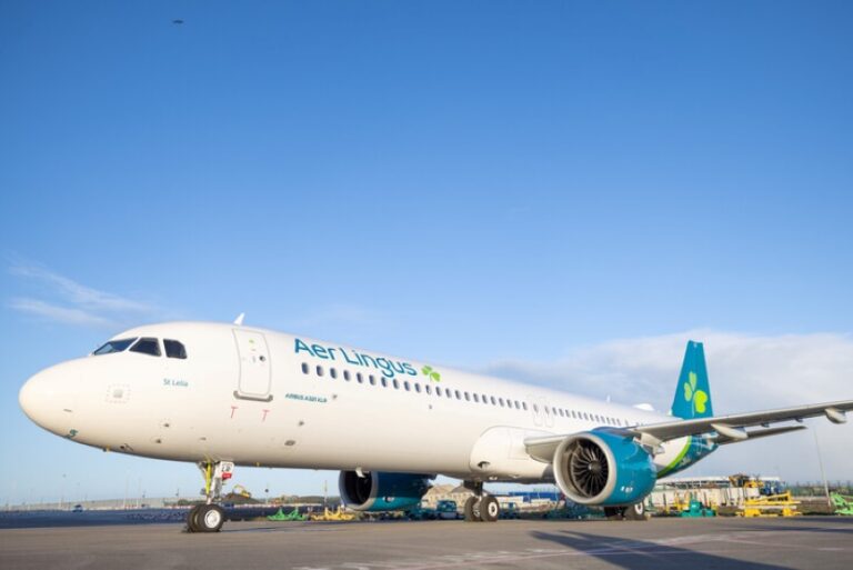 White Aer Lingus passenger jet parked on a sunny airport tarmac, teal tail with a shamrock logo visible under a clear blue sky.