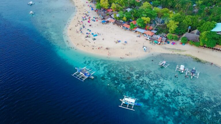 Aerial view of a tropical beach with white sand, colorful umbrellas, and huts along the shore amid green trees; clear turquoise water nearby.