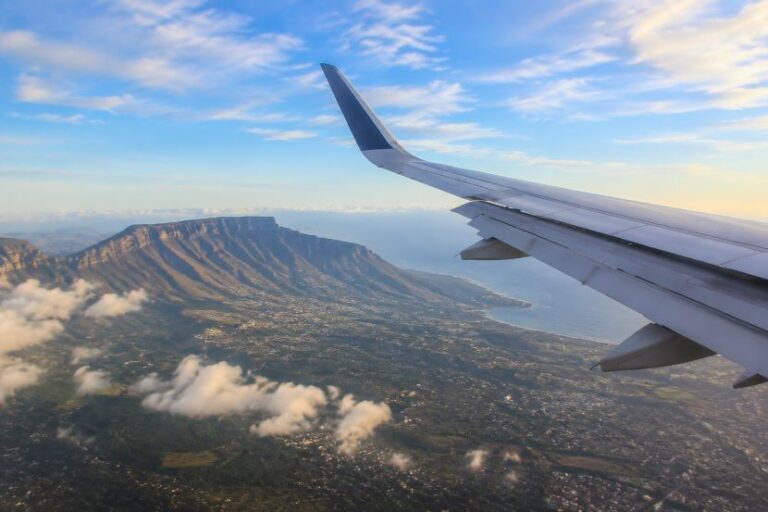 View from a plane window: wing in the foreground, coastline and rugged mountains below with scattered clouds in a blue sky.