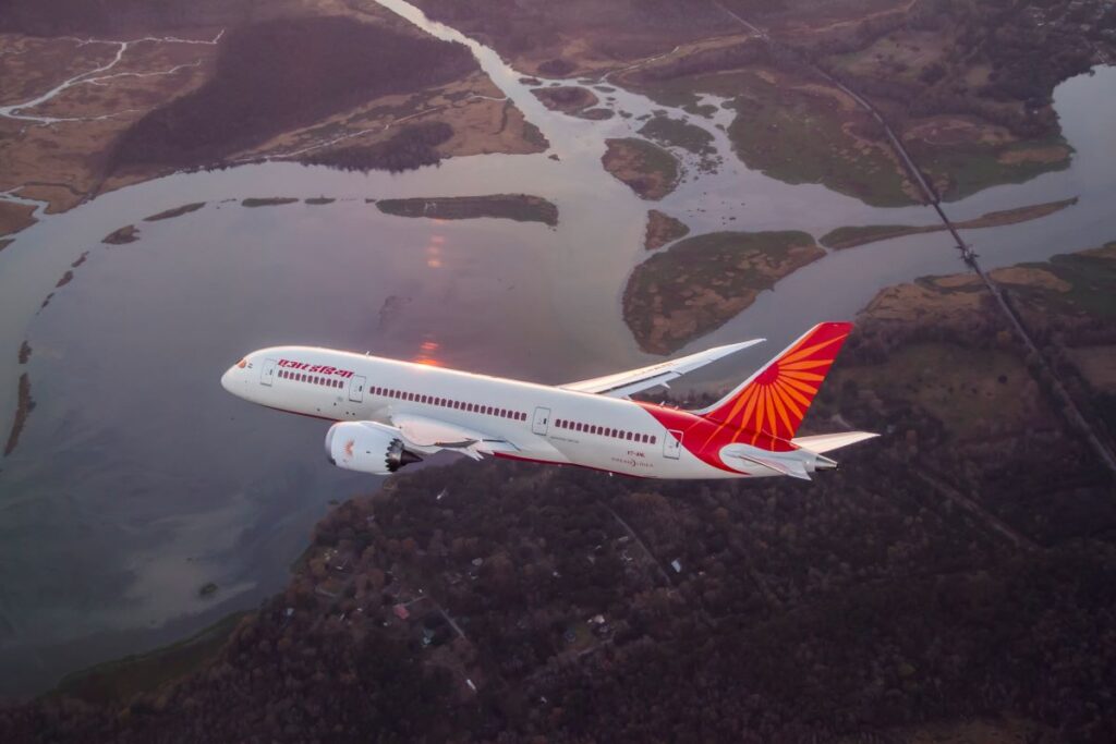 White passenger jet with a red-orange tail flying over a winding river and patchwork farmland below.