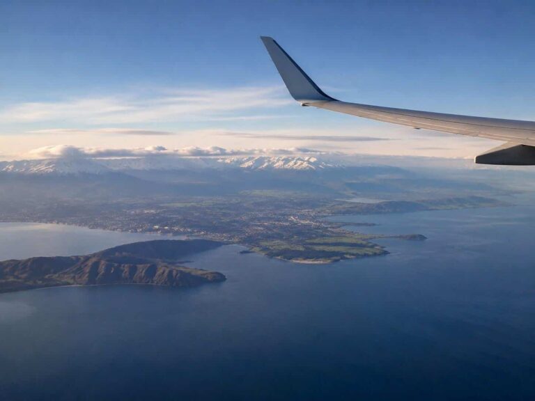 Airplane wing seen from a window over a coastal landscape with islands and snow-capped mountains below.