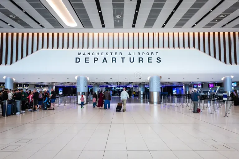 Manchester Airport departures hall with a large white sign reading 'DEPARTURES' and a crowd of travelers with luggage awaiting check-in/boarding.