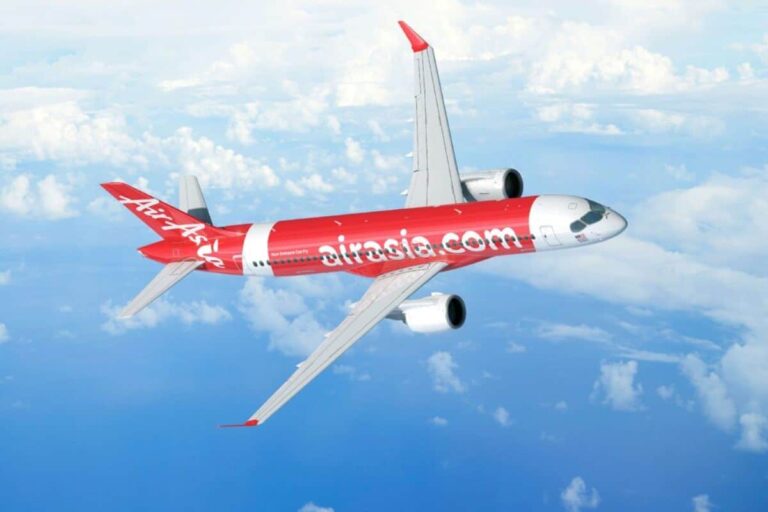 AirAsia jet with red fuselage and 'airasia.com' branding flying above clouds and blue sky.
