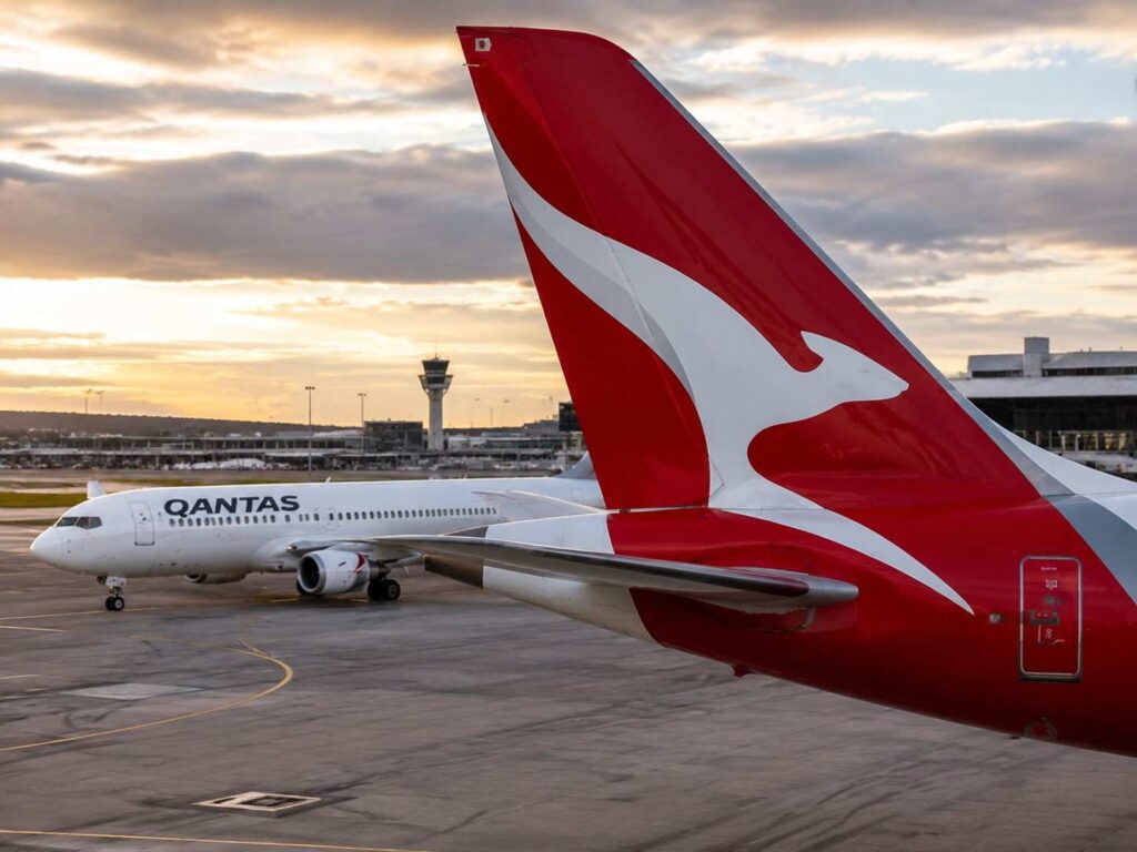 Qantas jet on the tarmac with a red-and-white tail logo at sunset, airport in the background