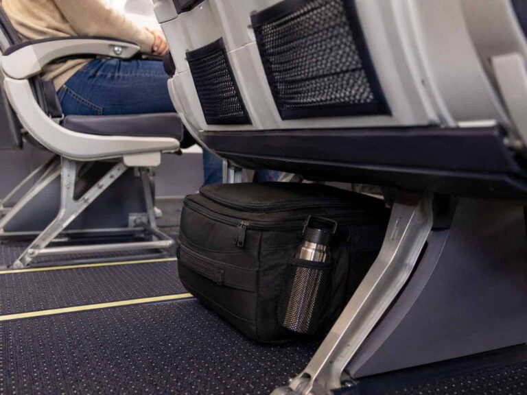 Black under-seat carry-on bag with a stainless steel water bottle in a mesh side pocket beneath an airplane seat.