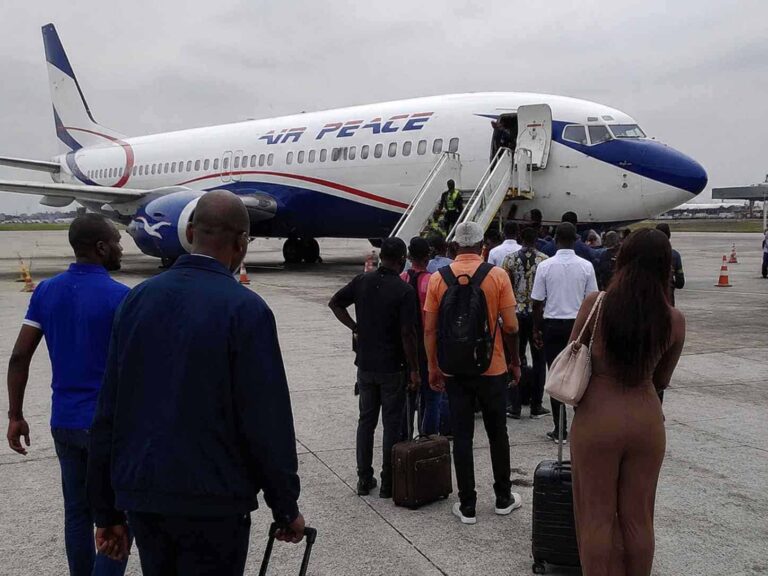 Line of passengers with rolling suitcases wait to board a white Air Peace aircraft via mobile stairs on the tarmac.