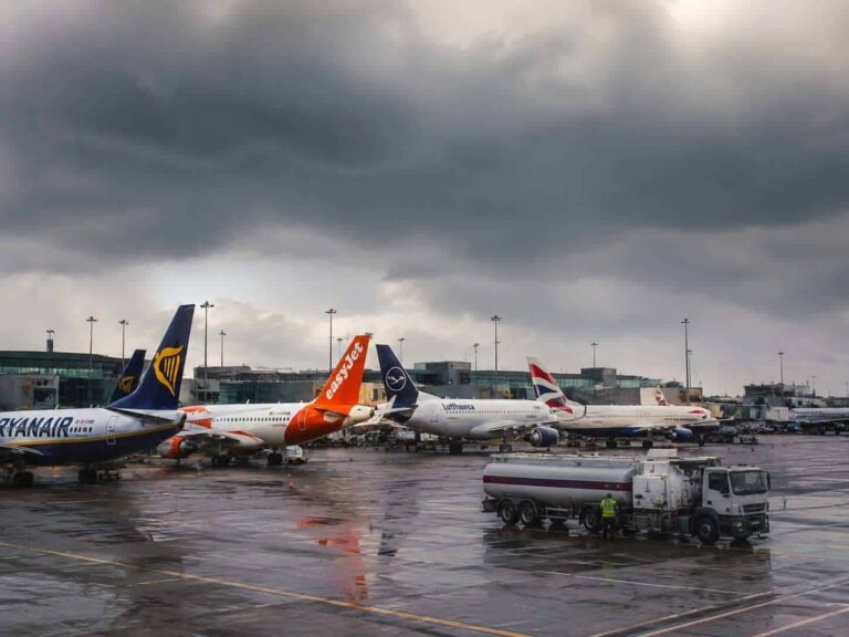 Row of parked commercial jets on a wet tarmac beneath dark storm clouds, with a fuel truck in the foreground.