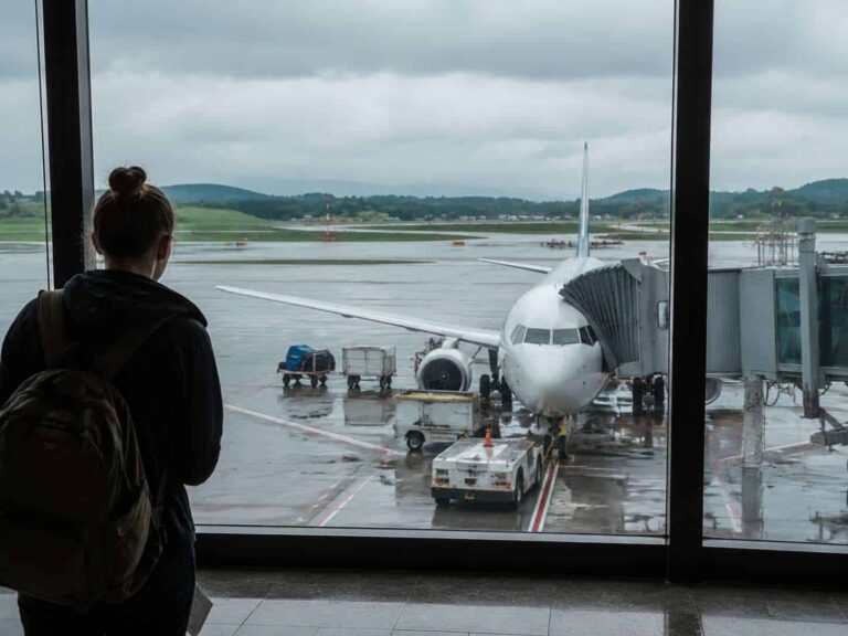 Traveler with a backpack stands at the window, watching a white airplane at the gate on a wet tarmac.