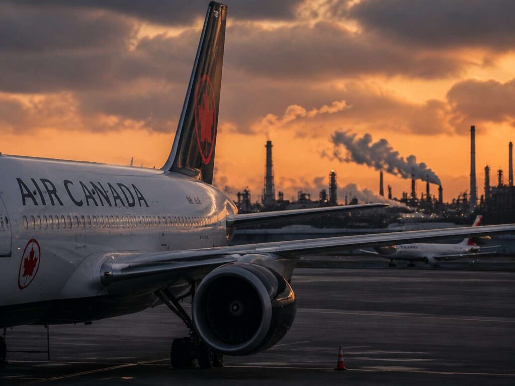 Air Canada jet on the taxiway at sunset with an industrial skyline and smokestacks in the distance.
