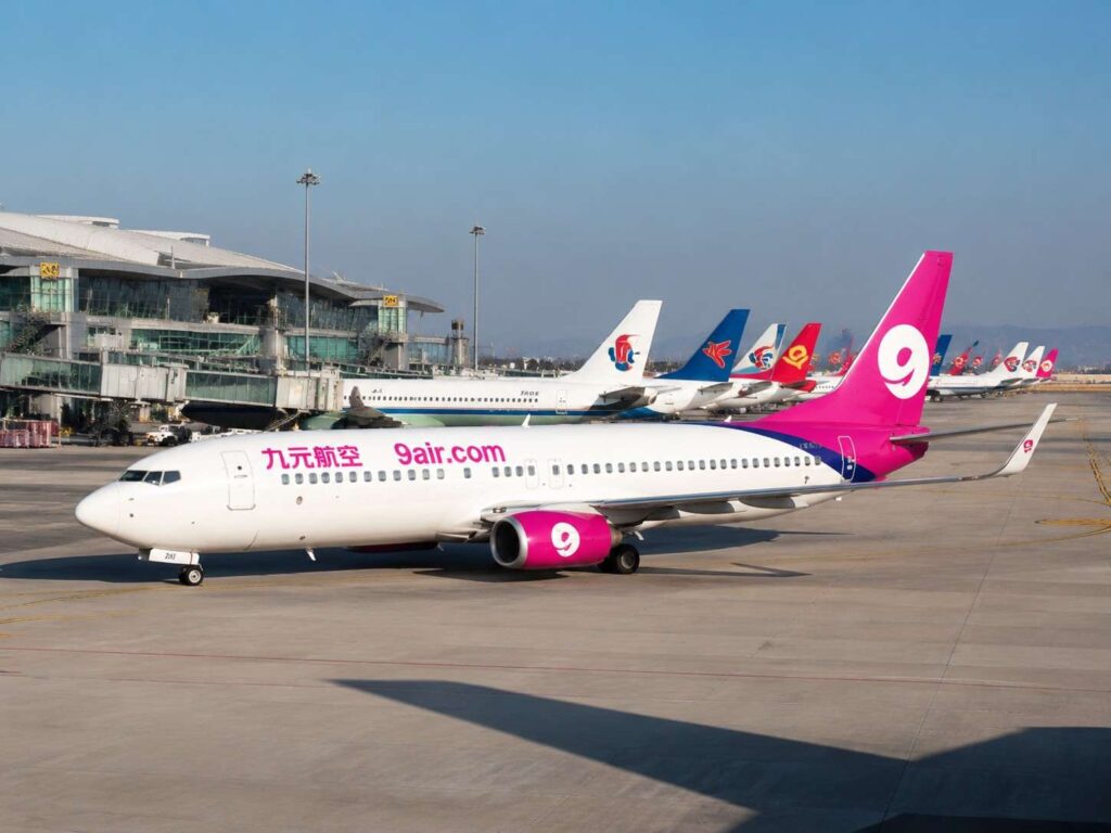 White passenger jet with pink tail and 9air.com branding parked on the tarmac at an airport, with terminal and other planes in the background.