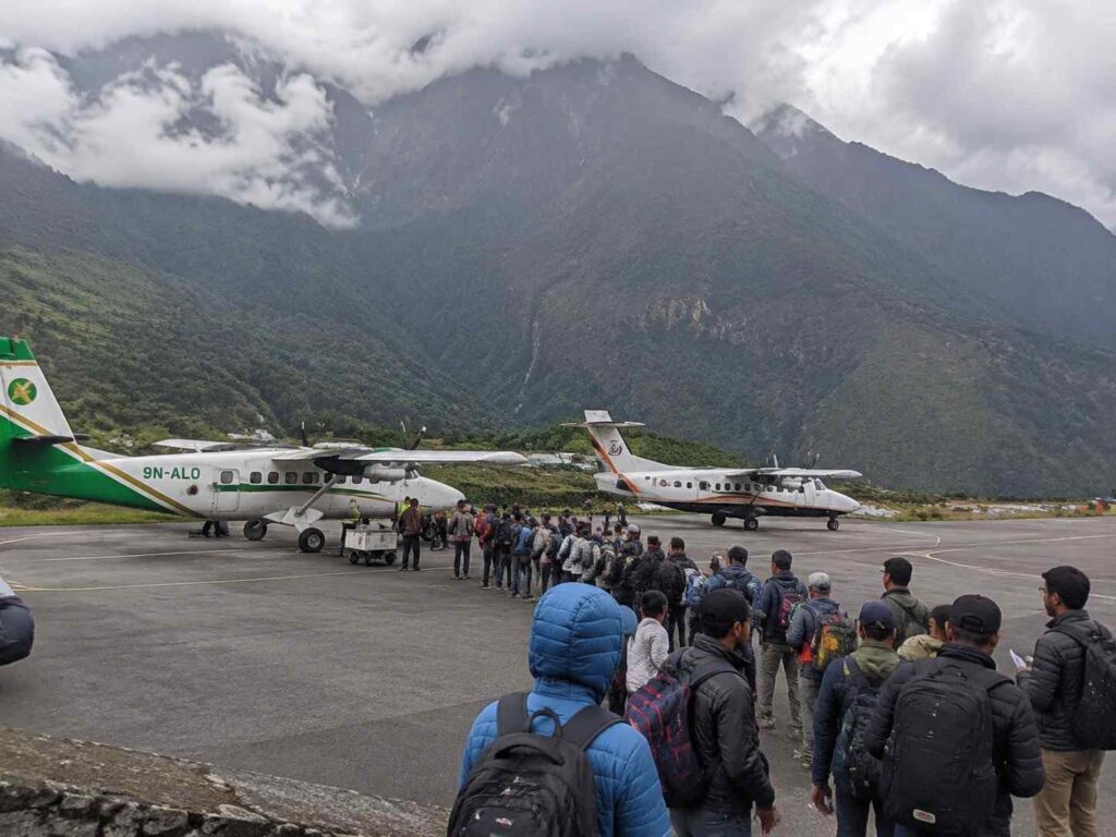 Line of travelers with backpacks waiting to board two small propeller planes at a mountain airstrip.